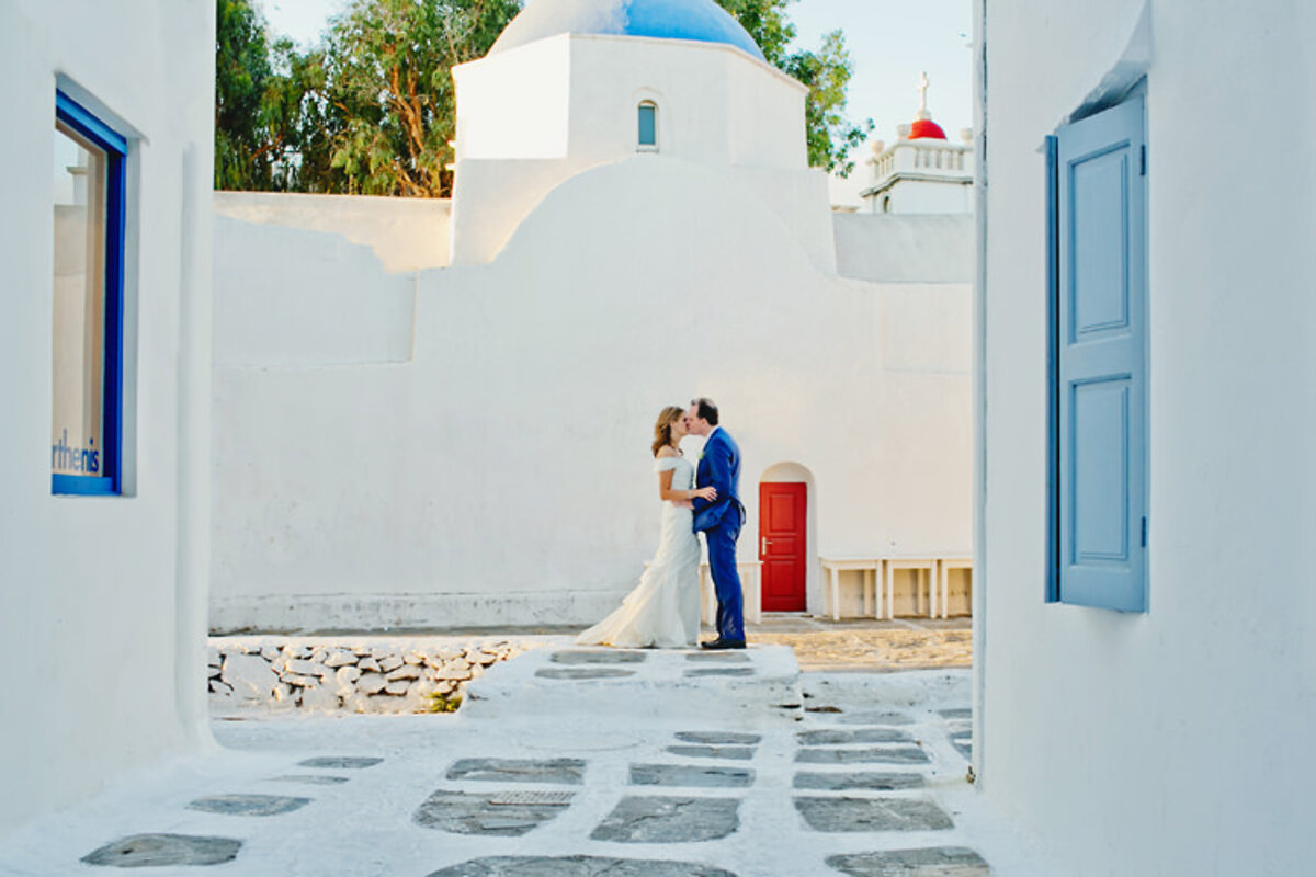 A couple kissing in front of Mykonos Catholic church beautifully documenting wedding love stories in Greece's picturesque setting. Mykonos catholic ceremony packages