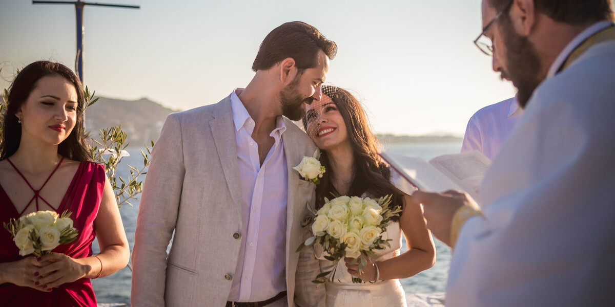 A romantic Greek Orthodox wedding ceremony by the sea in Mykonos, with the bride and groom sharing a joyful moment as the priest officiates