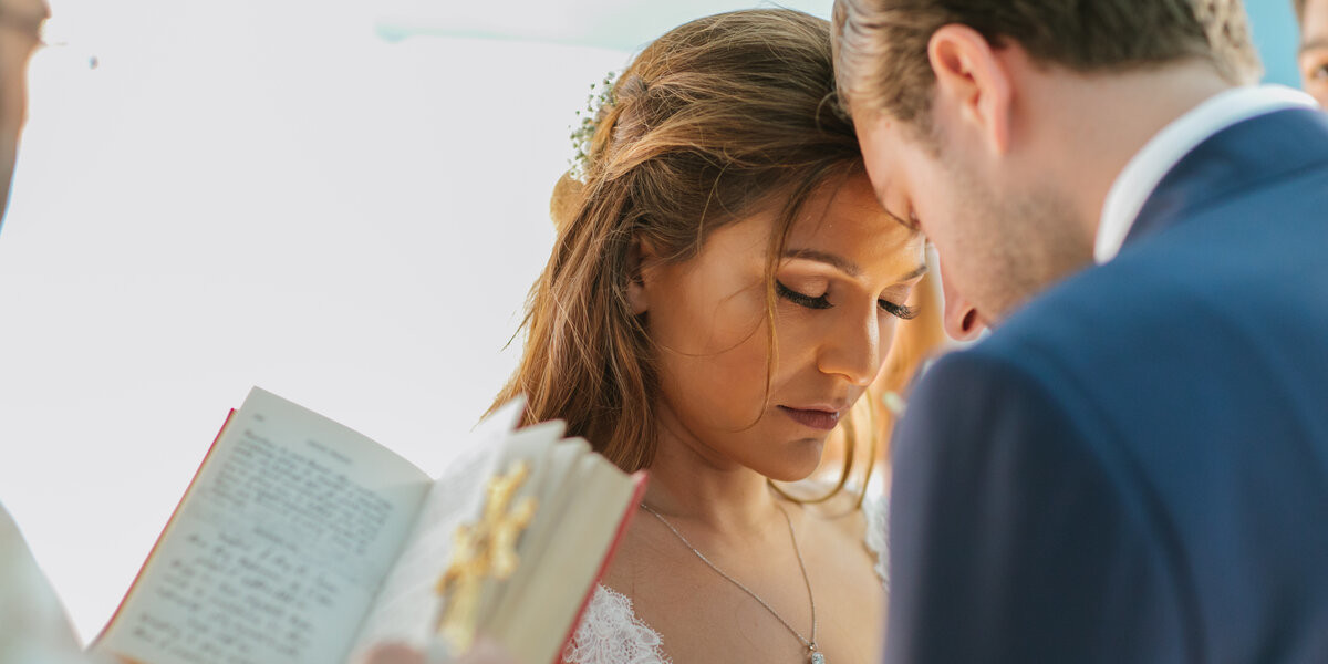 A deeply emotional moment during a Greek Orthodox wedding ceremony, as the bride and groom bow their heads in prayer, with a priest holding a religious book and cross.