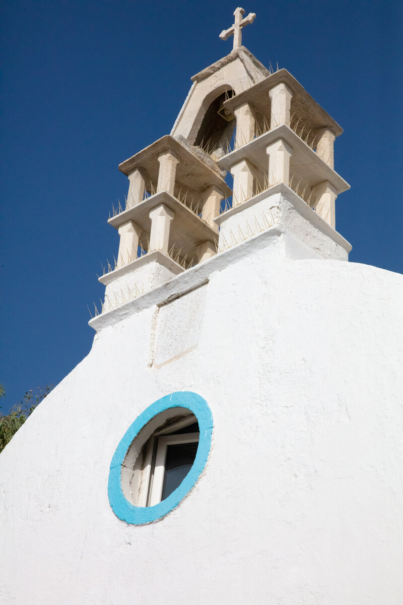 White Mykonos catholic chapel with a blue circular window and a distinctive bell tower topped with a cross against a clear blue sky.