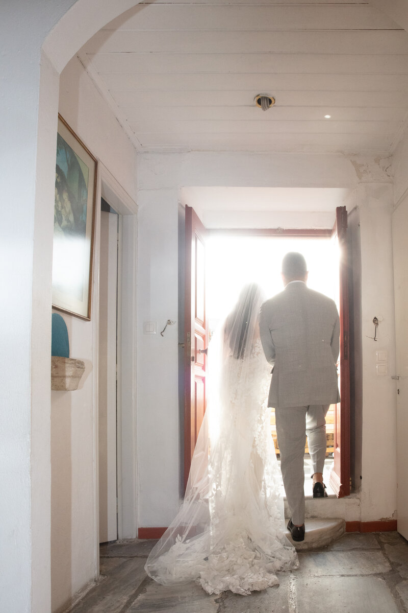 A bride in a flowing white wedding gown and veil stands beside her groom, both silhouetted against the bright doorway of the Catholic church in Mykonos, ready to step outside.